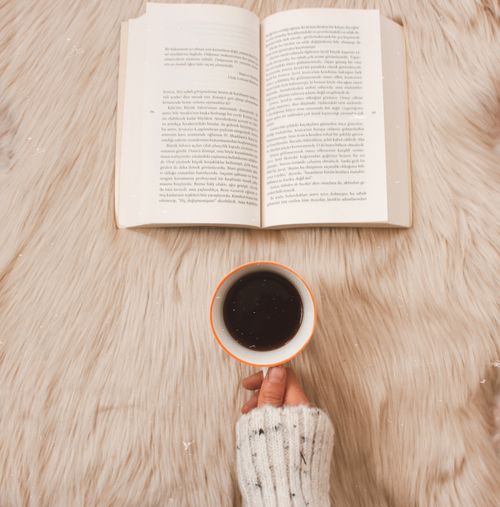 Free A cozy scene with a hand holding a coffee cup and an open book on a soft rug. Stock Photo