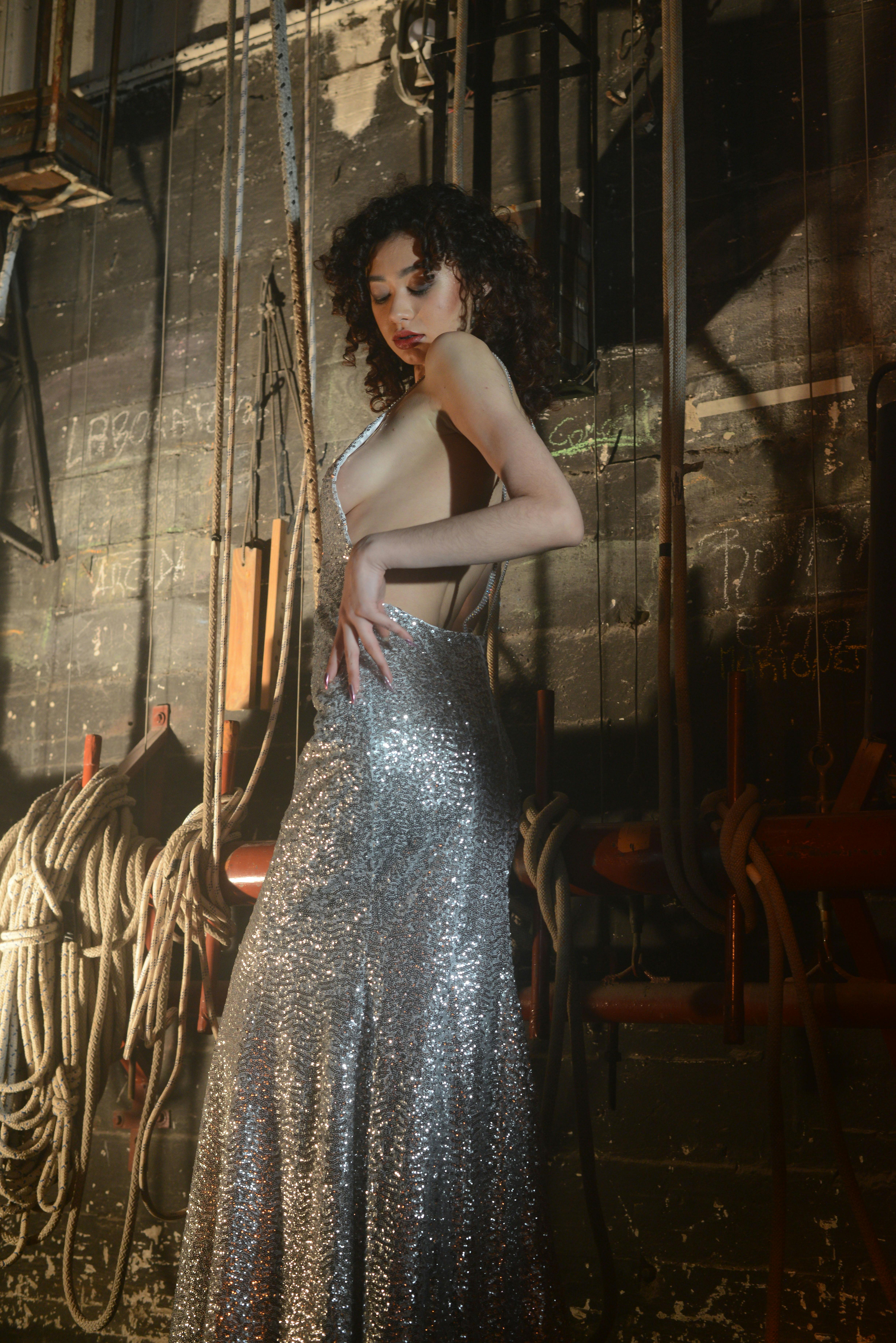 Free An elegant woman in a sparkling silver dress poses on a dramatic stage set with ropes. Stock Photo