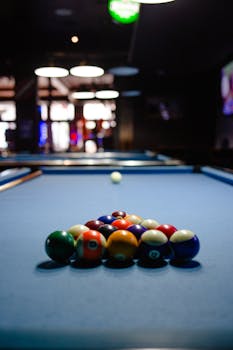 Colorful billiard balls arranged on a pool table in a dimly lit game room setting.