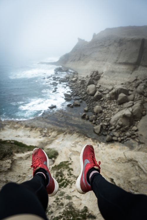 Free Experience adventure sitting at the edge of a cliff overlooking a dramatic rocky coastline and roaring ocean waves. Stock Photo