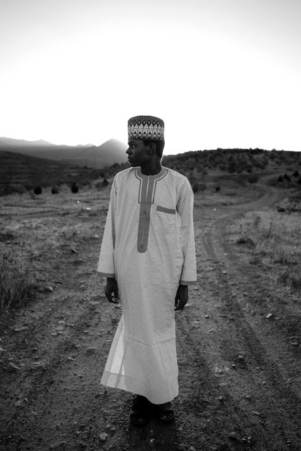 Black and white photo of a man in traditional attire standing on a rural dirt road.