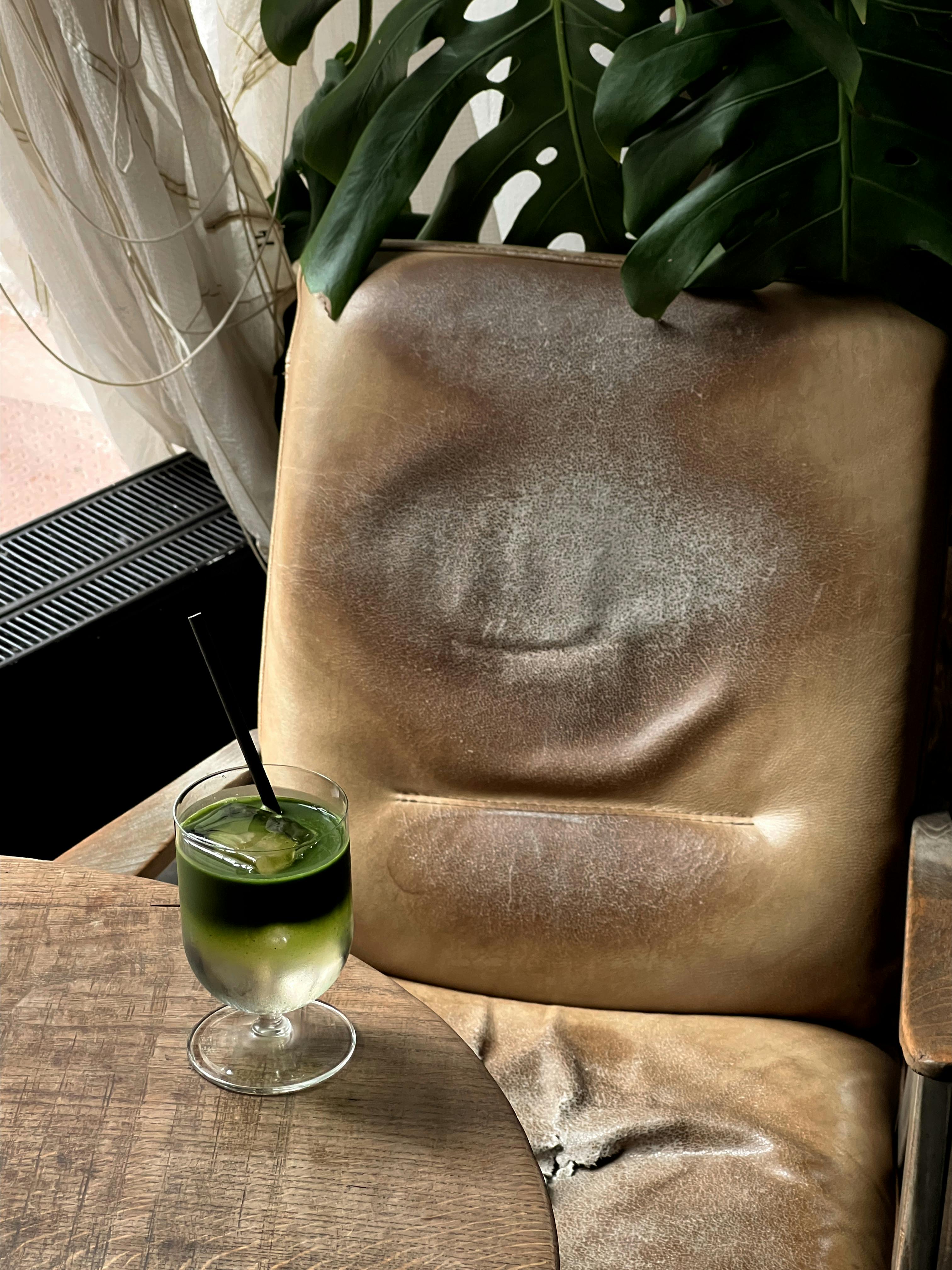 A green cold drink on a wooden table beside an old leather chair with green plants.