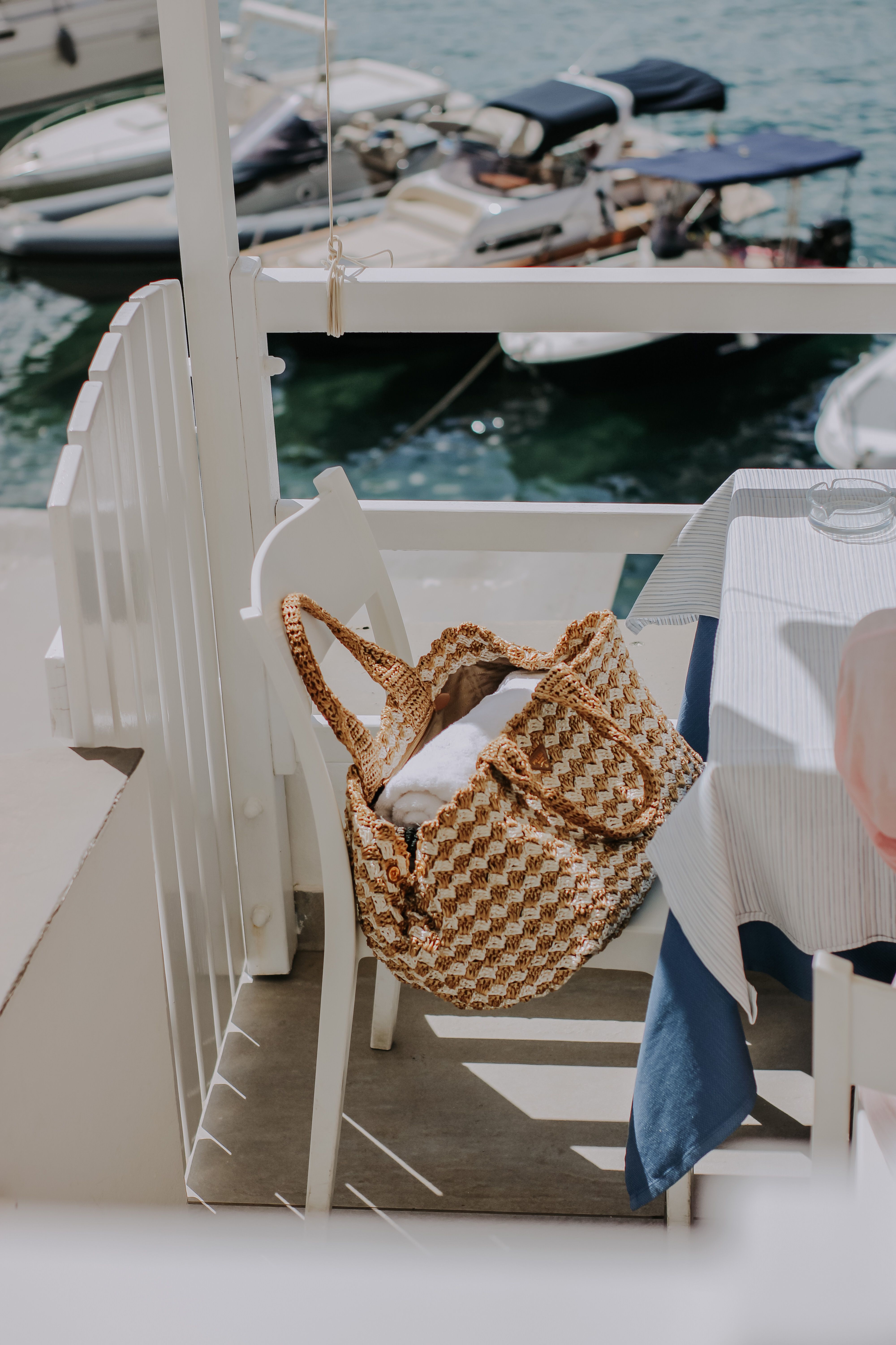 Woven bag on a sunlit patio with boats in a scenic harbor view.