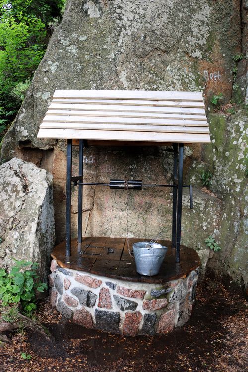 Free A rustic stone well with a metal bucket, set against a rocky backdrop in a rural setting. Stock Photo