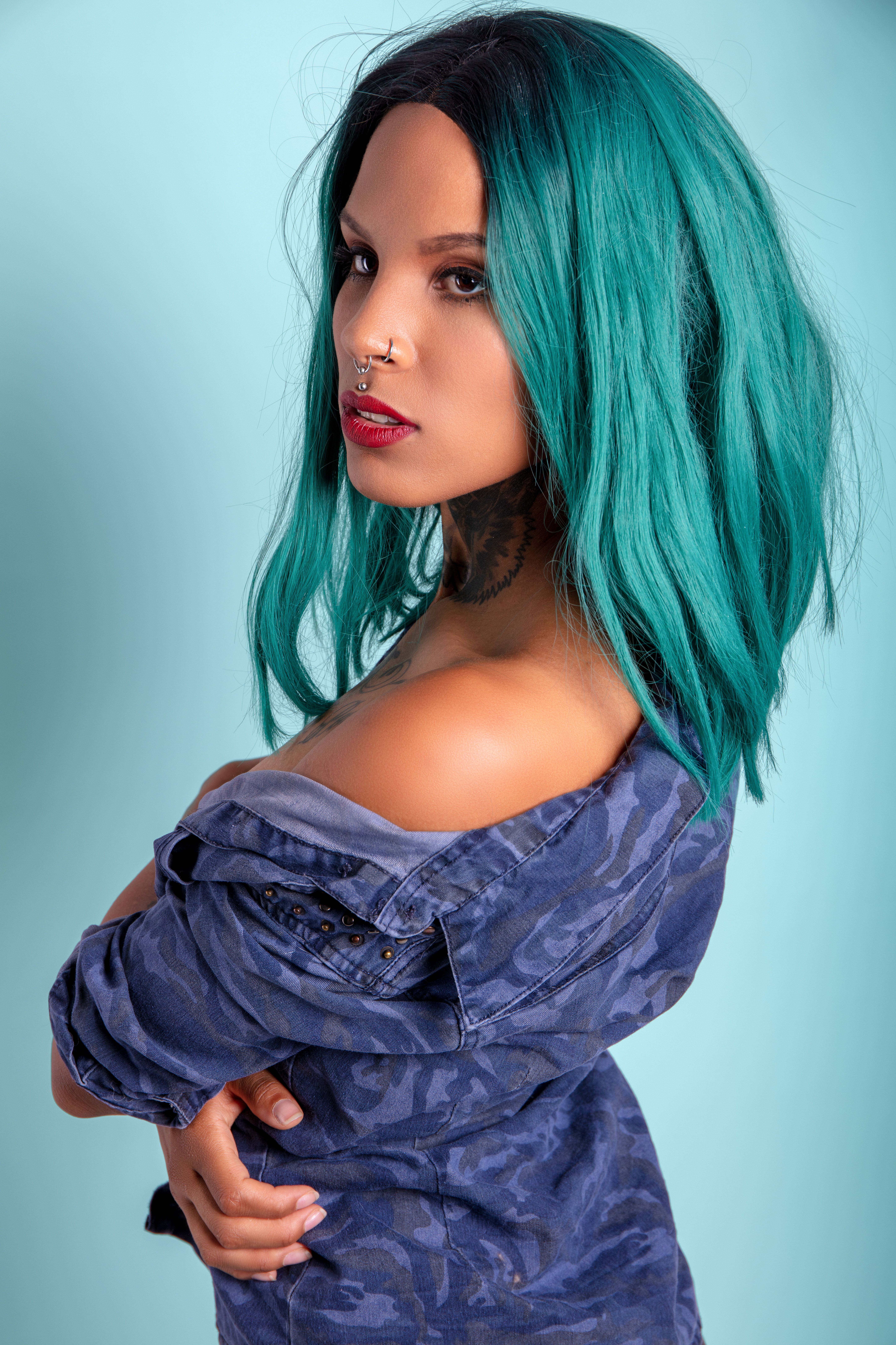 Stylish portrait of a woman with vibrant blue hair, piercings, and tattoos, wearing off-shoulder denim against a blue backdrop.