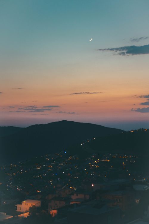 Free Peaceful mountain town at twilight with a crescent moon and city lights below. Stock Photo