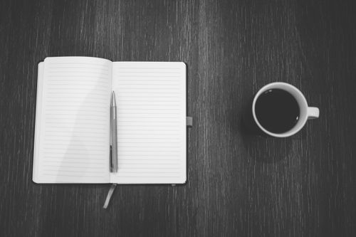 Free Black and white shot of a notepad and coffee cup on a wooden table from above. Stock Photo
