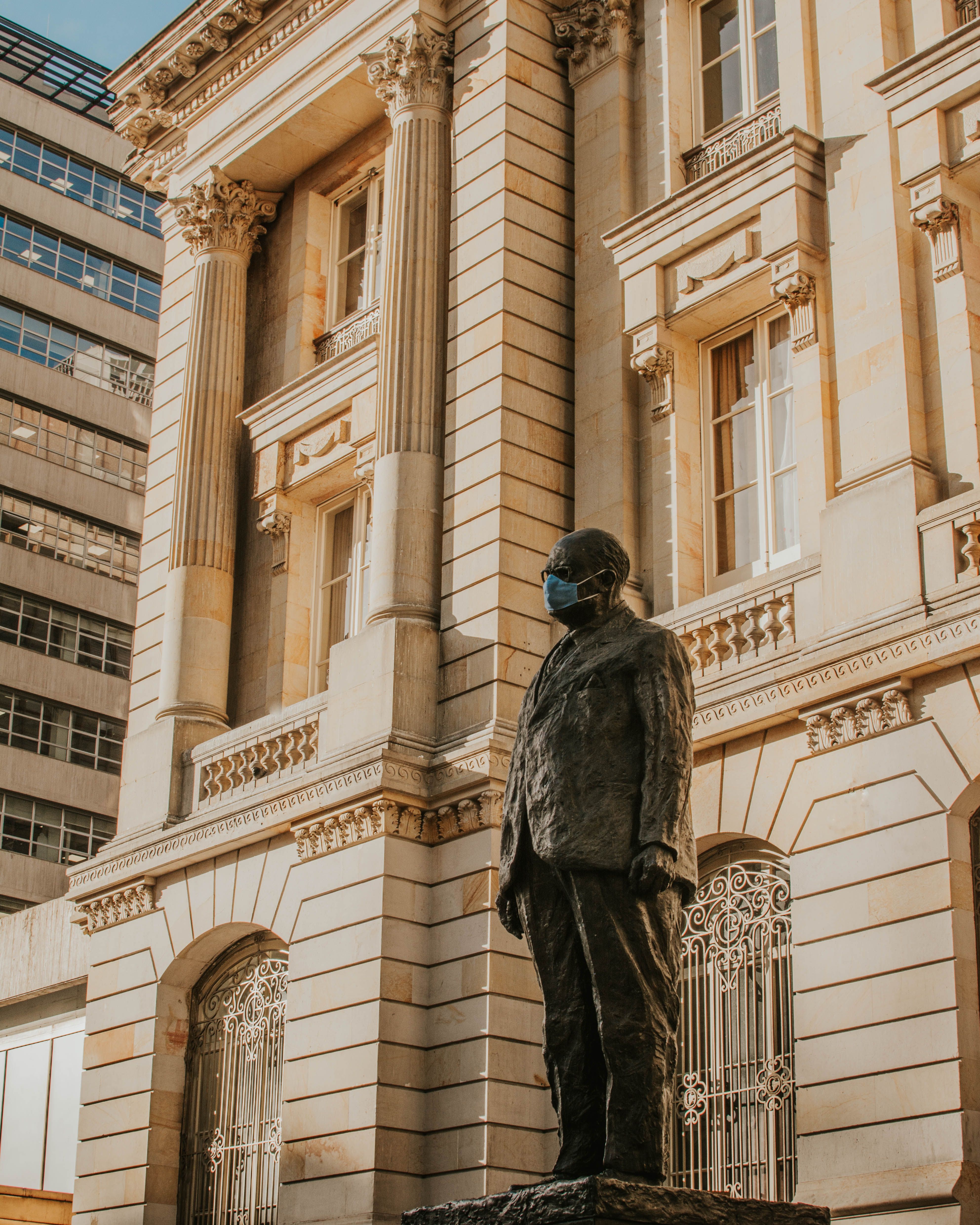 A statue of Alfonso Lopez Michelsen wearing a face mask in front of a neoclassical building in Bogotá, Colombia.