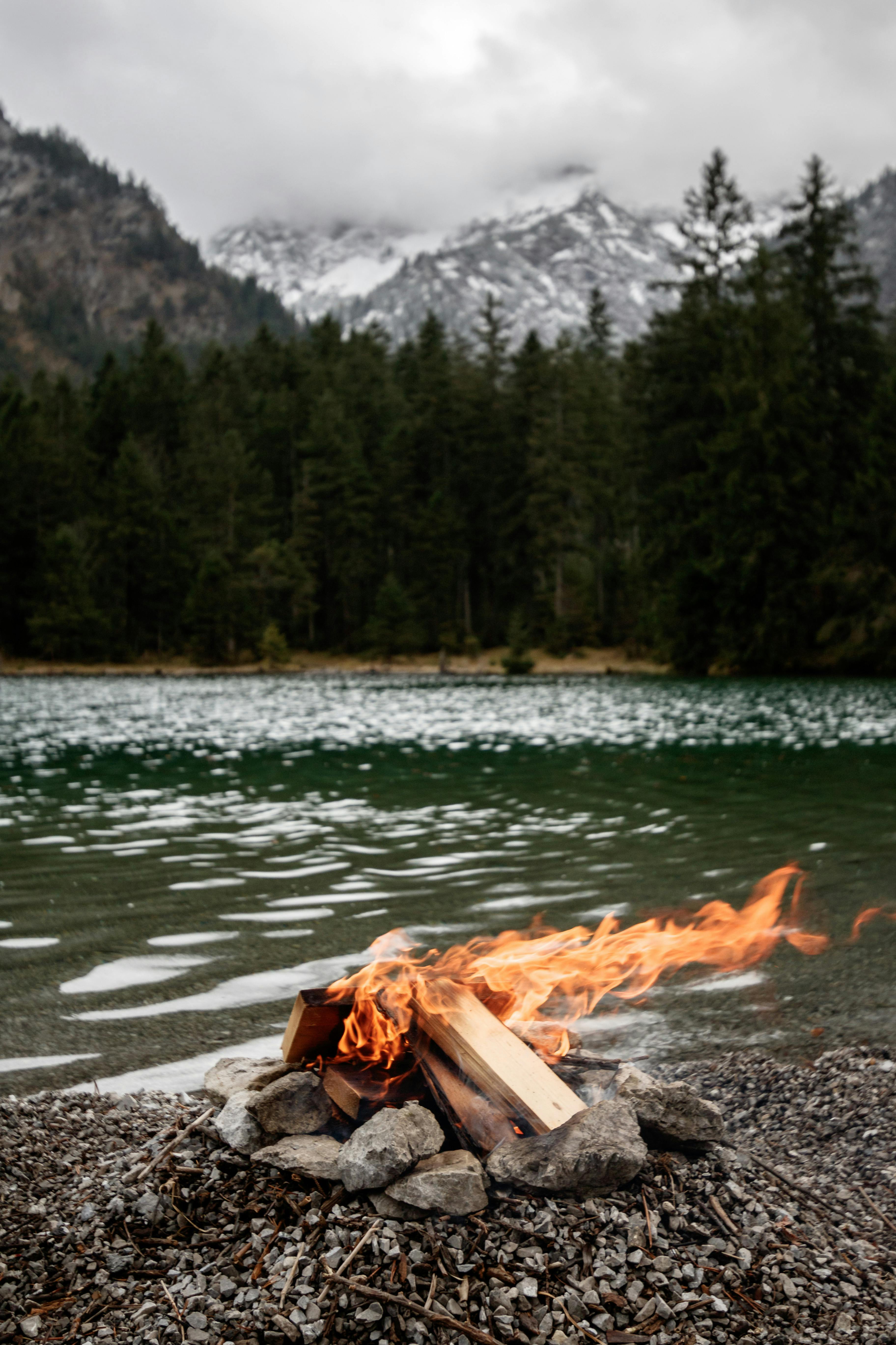 Cozy bonfire by a serene mountain lake with snowy peaks in the background.
