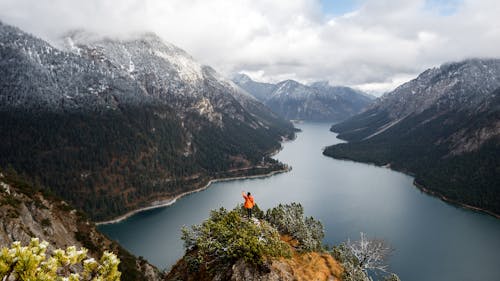 A Person in Orange Jacket Standing on Rock Near Lake