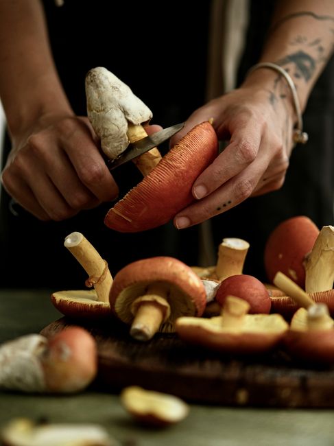 Tattooed hands expertly slicing fresh mushrooms on a wooden board in a kitchen setting, emphasizing culinary skill.