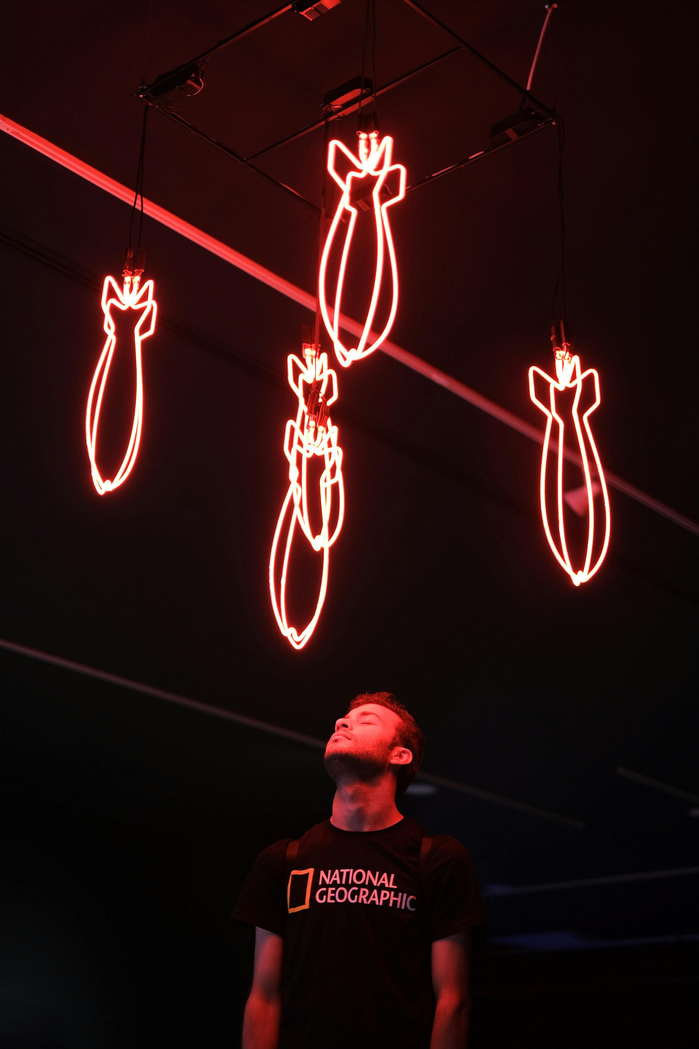 Man standing under red neon bomb lights in a dark room, looking upwards pensively.