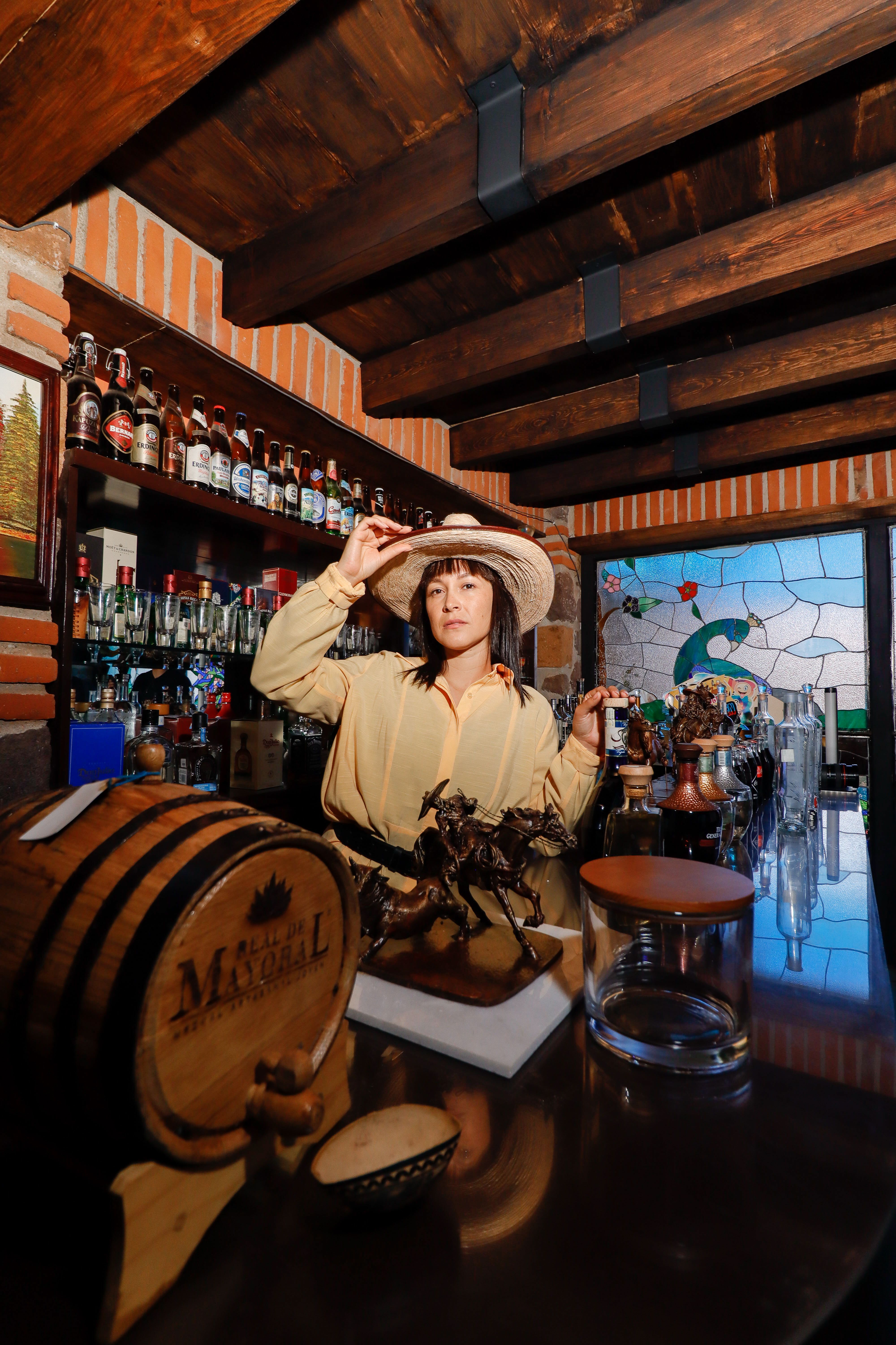 Elegant woman in a hat posing in a traditional Mexican bar with rustic wooden decor.