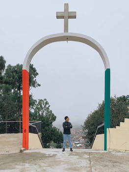 A person poses under a red, white, and green archway with a cross on a cloudy day.