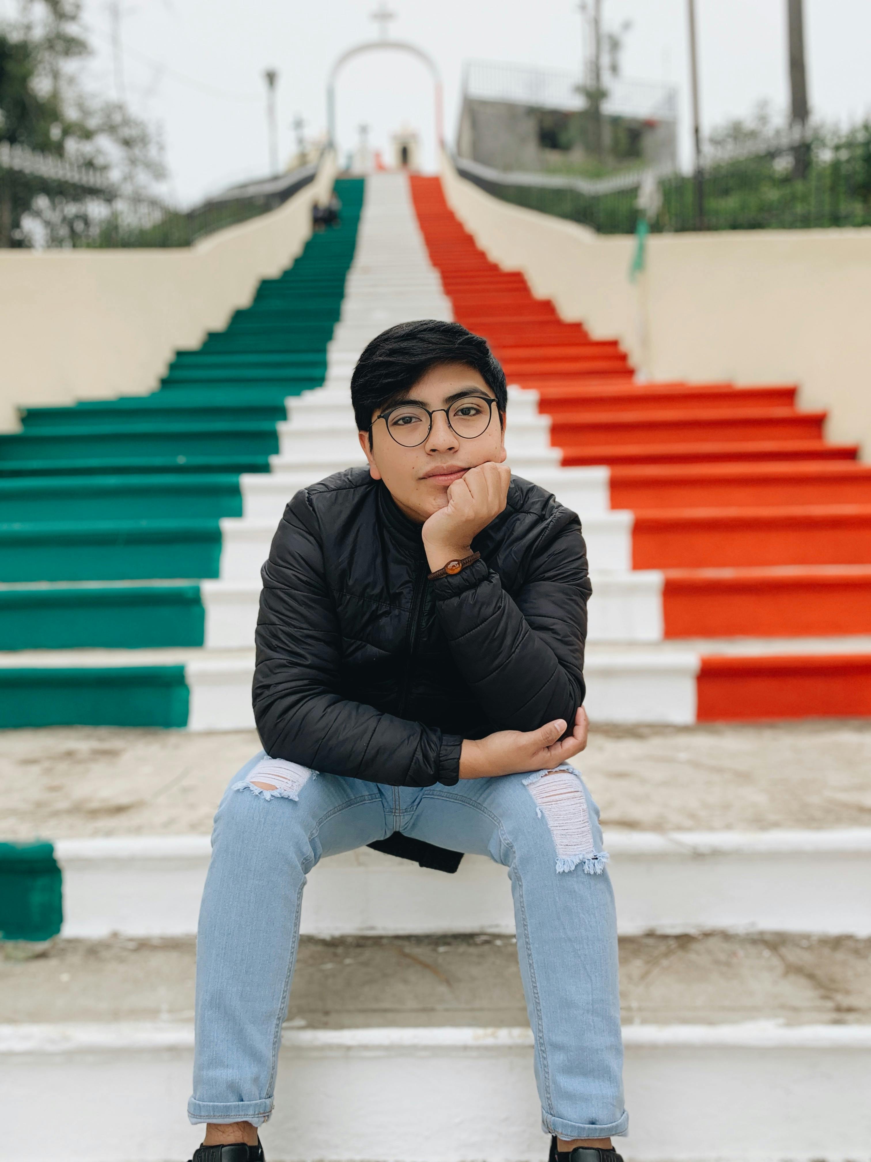 Young man in a leather jacket sits on steps painted with vibrant colors in San Andrés Larráinzar, Mexico.