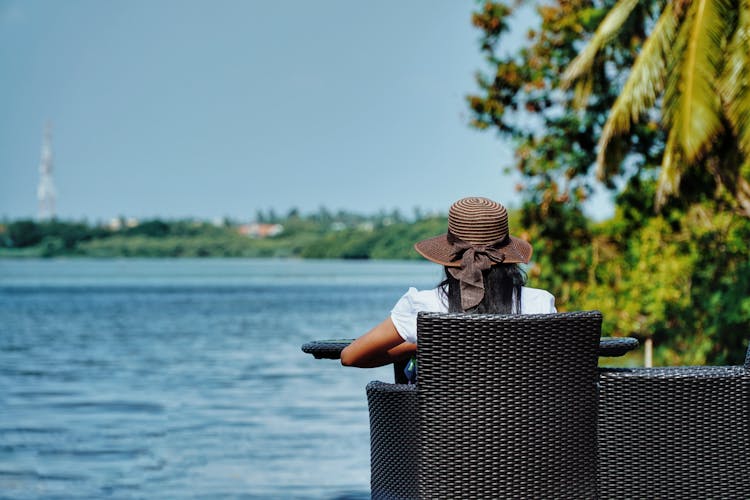 Person Wearing Sun Hat Sitting On Rattan Garden Chair In Front Of A Sea