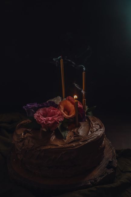 Dark chocolate cake adorned with elegant flowers and candles glowing softly in a moody setting.