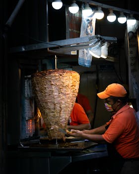 A street vendor prepares shawarma kebabs under bright lights, carving meat from a vertical rotisserie.