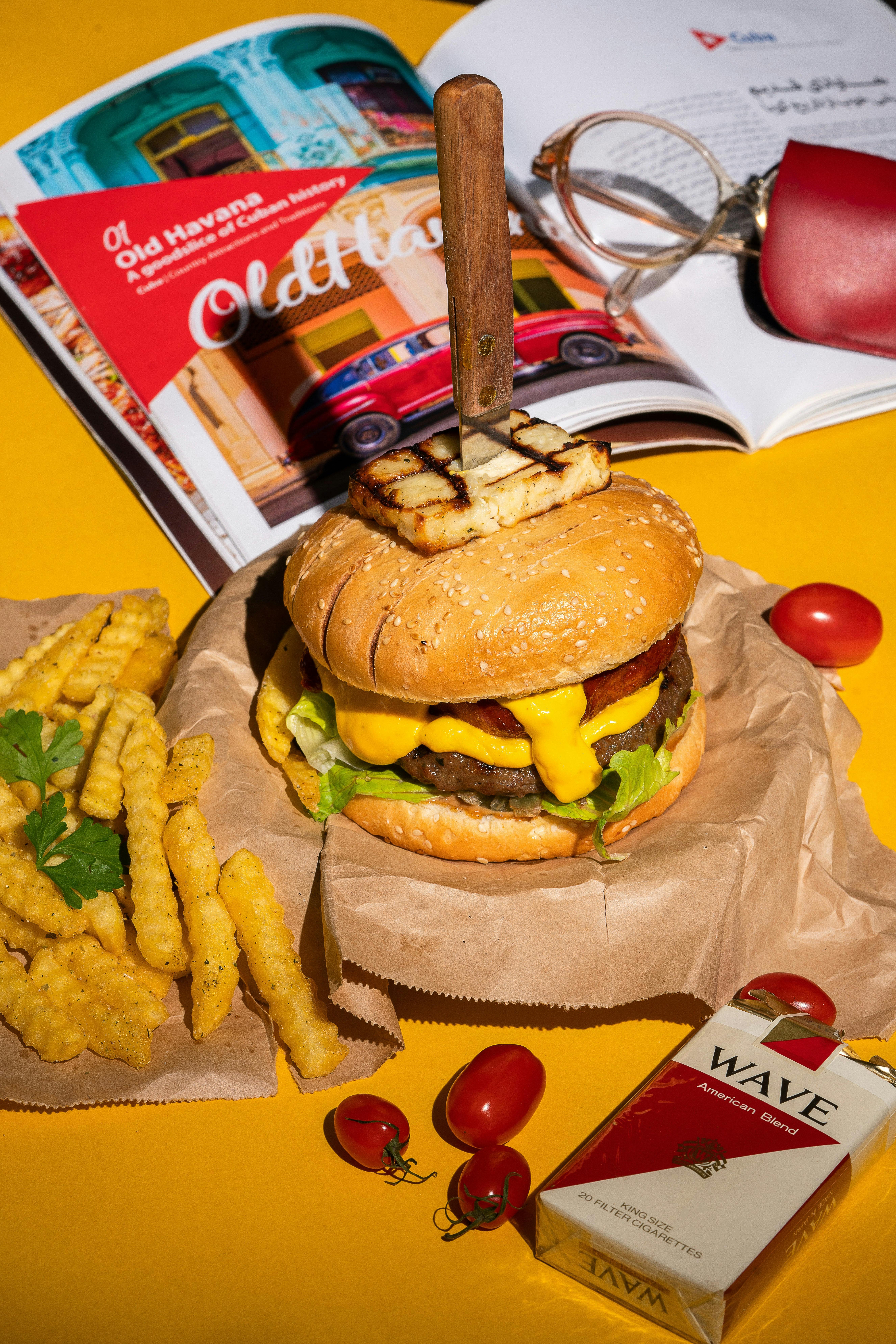 Delicious gourmet burger with crinkle-cut fries on yellow surface, styled with magazines and accessories.