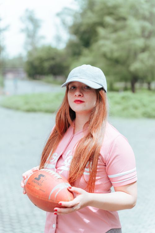 Free A woman in a pink shirt and cap holds a football outdoors, exuding confidence. Stock Photo