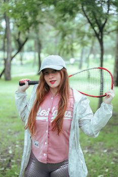 A young woman with long hair and a raincoat holds a badminton racket in a lush green park.