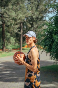 Athletic young man in a cap and tank top holding a basketball outdoors in a park setting.