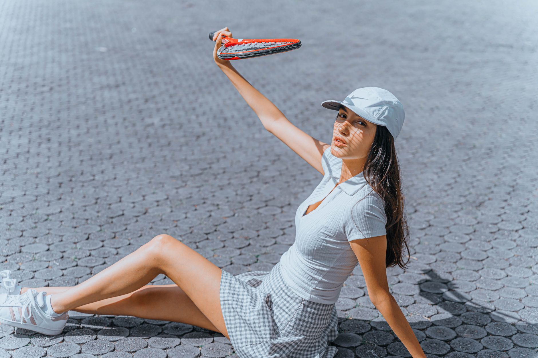 Stylish woman in a checkered skirt holds a tennis racket while sitting on pavement outdoors.