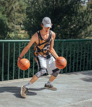 A young man outdoors practicing basketball dribbling with two balls in an athletic outfit.