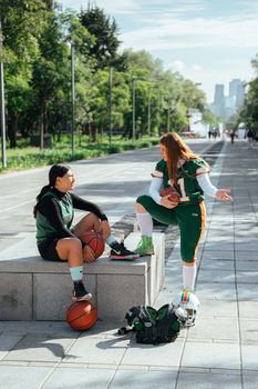 Two women athletes discussing on a pavement with sports equipment outdoors.