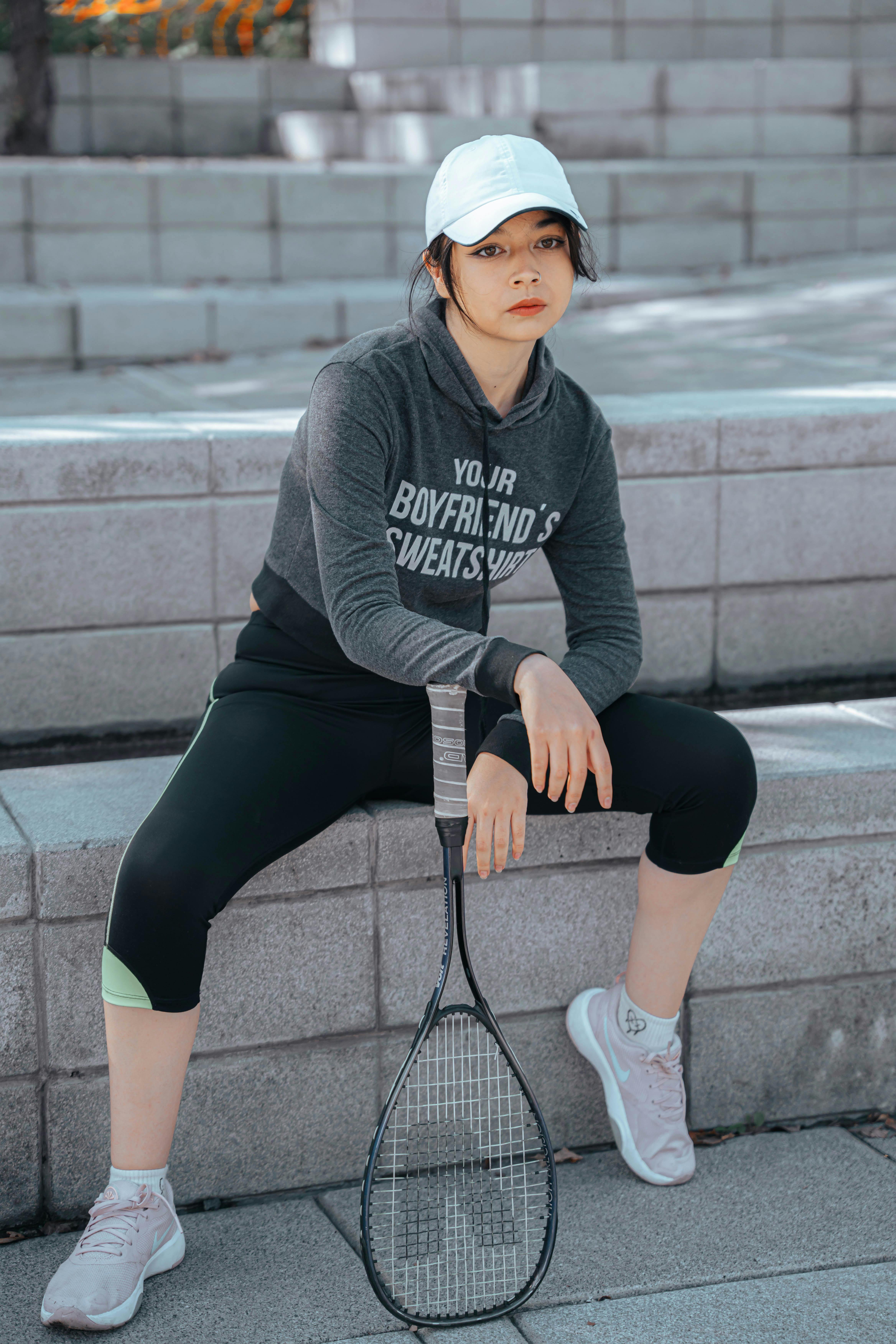Woman in sportswear sits confidently on steps holding a squash racket outdoors.