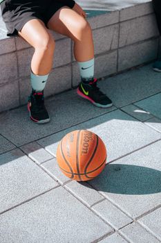 A basketball rests outdoors on a sunny day with a player's legs in the background.
