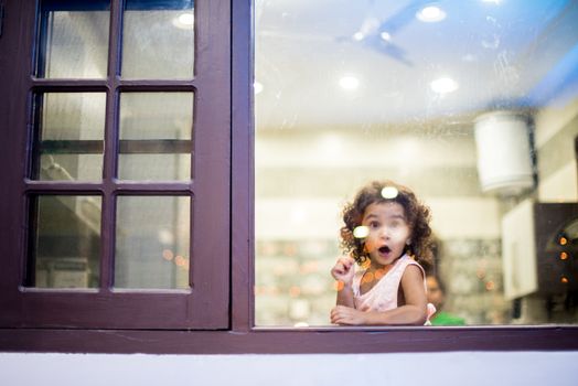 A child with a surprised expression peers through a glass window indoors in India.