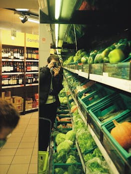 Man taking a selfie in a supermarket aisle with vegetables and wine displayed.