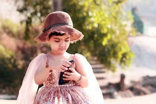 Free A young girl wearing a brown hat and sequin dress uses a smartphone outdoors. Stock Photo