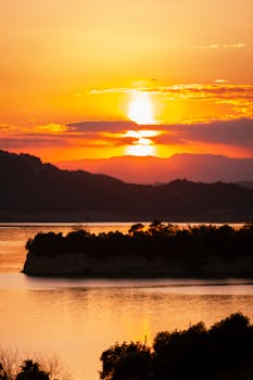 Stunning sunset over a tranquil river with silhouettes of hills and lush vegetation.