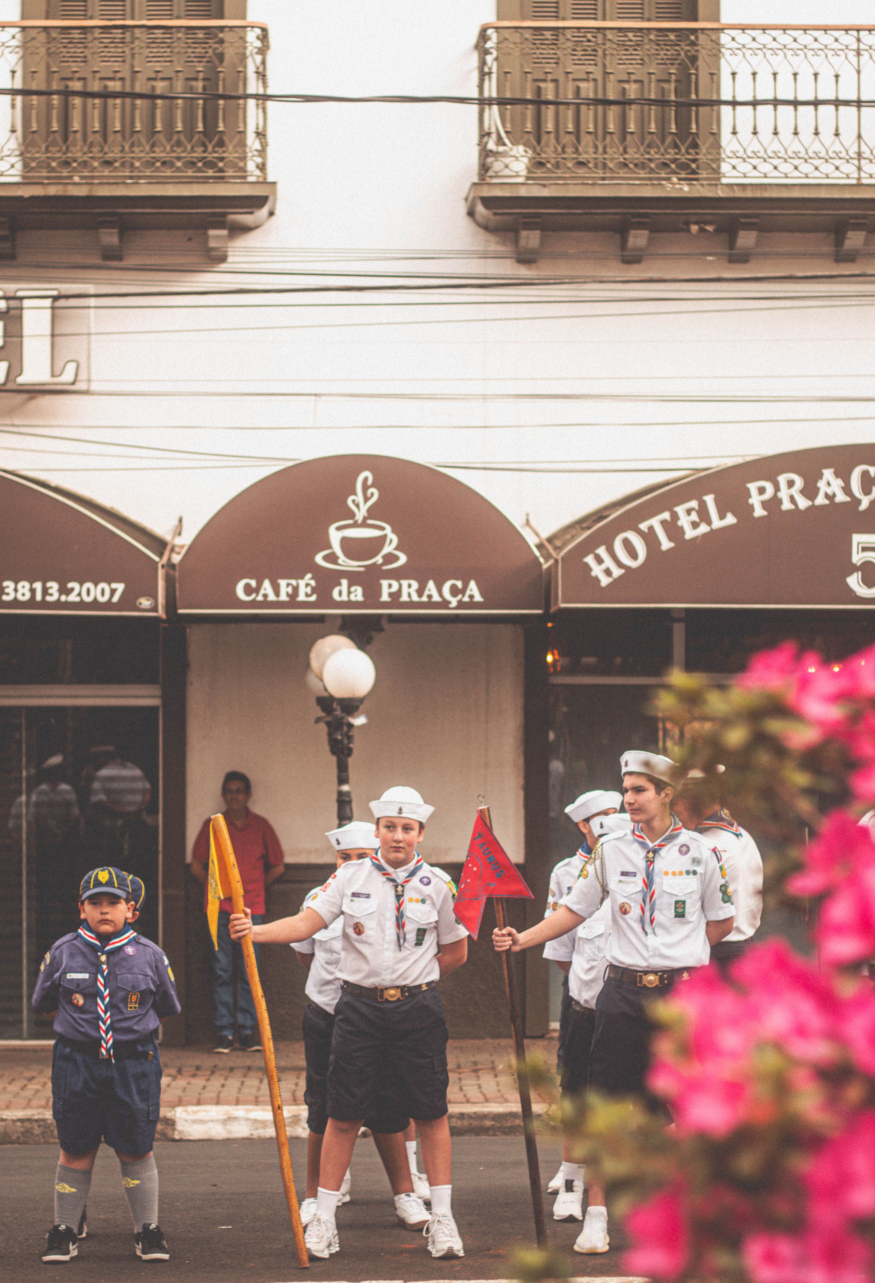 Youth scouts in uniform standing in formation on a city street outside a cafe and hotel.