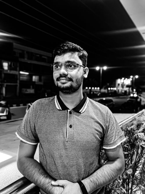 Free Grayscale portrait of a man with a beard wearing eyeglasses, standing on a city street at night. Stock Photo