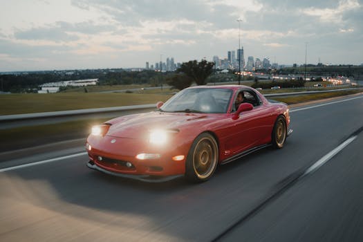 A red Mazda RX-7 sports car driving on a highway with Calgary skyline in the background.