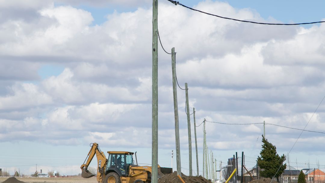 A backhoe digging at a site beneath utility poles and overhead wires