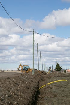 A backhoe digging at a construction site with utility poles and overhead wires under a cloudy sky.