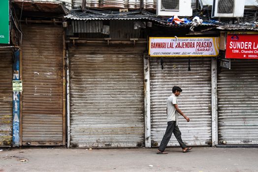 A man walks past closed metal shutters on an urban street with shop signs overhead.