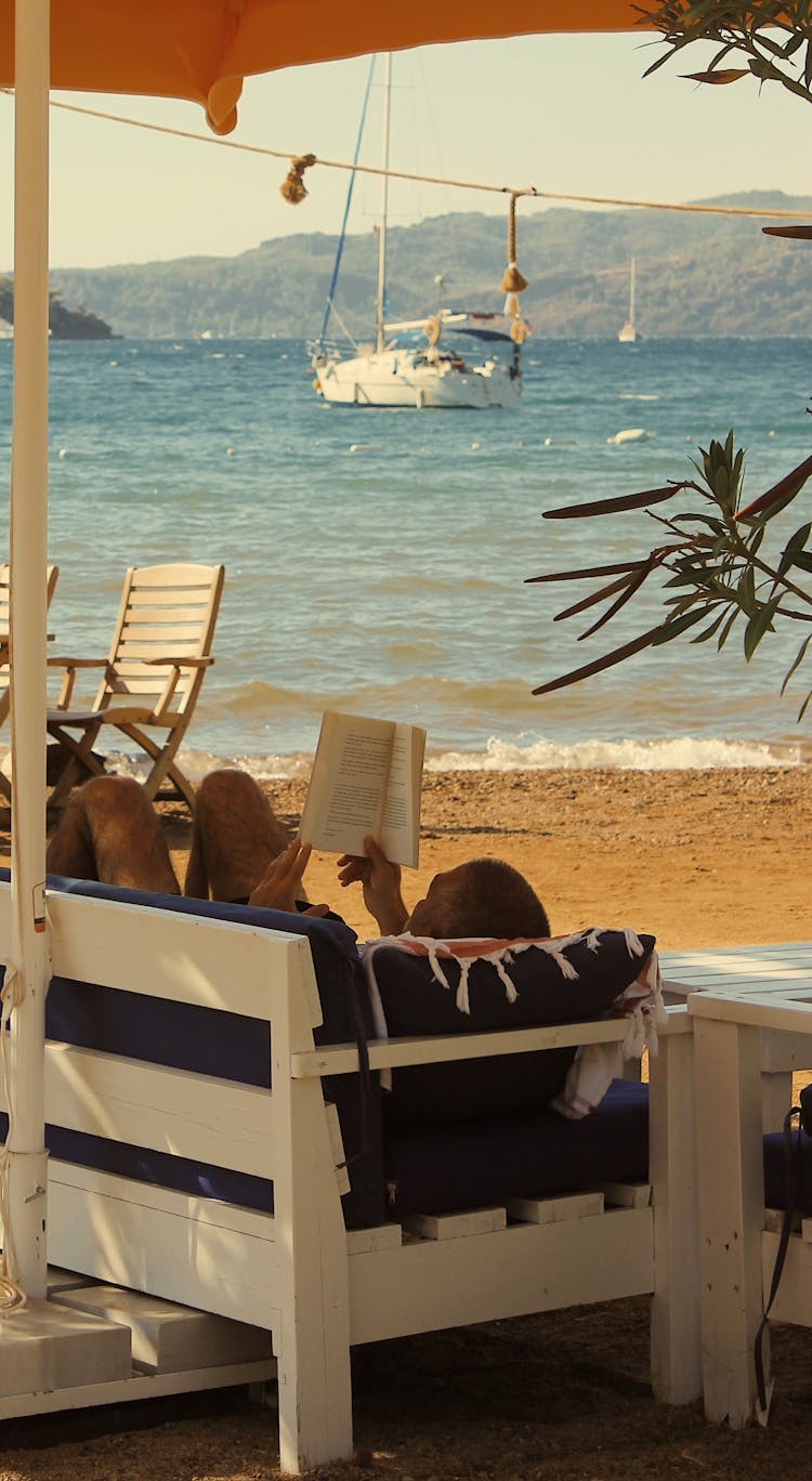 A Man Lying On A Wooden Lounge While Reading A Book