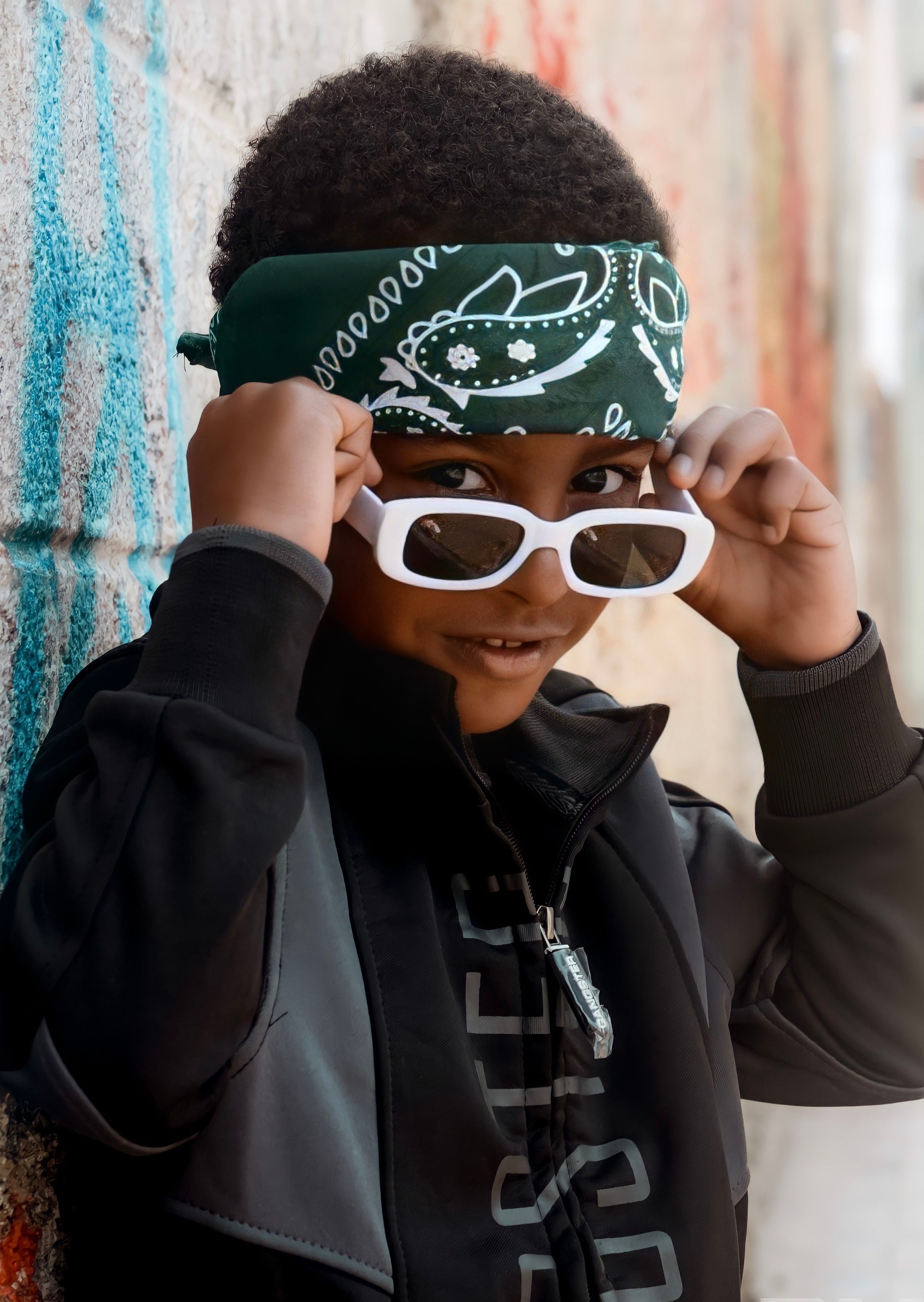 A stylish young boy in a bandana and sunglasses poses against a vibrant wall in São Paulo, Brazil.