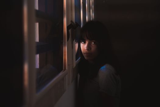 A woman leans by a window in a dark room, illuminated by soft natural light.