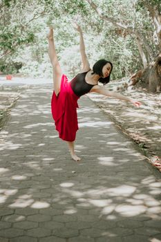 Asian woman in a red skirt performs ballet on a sunlit forest path.
