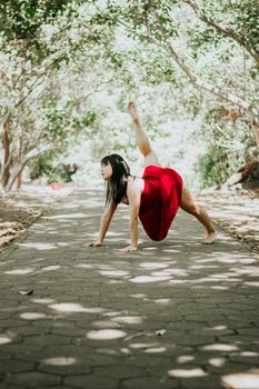 A woman in a red dress performs a dance pose surrounded by lush greenery on a sunlit path.