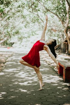 A woman in a red dress dancing barefoot on a tree-lined path in a sunny park.