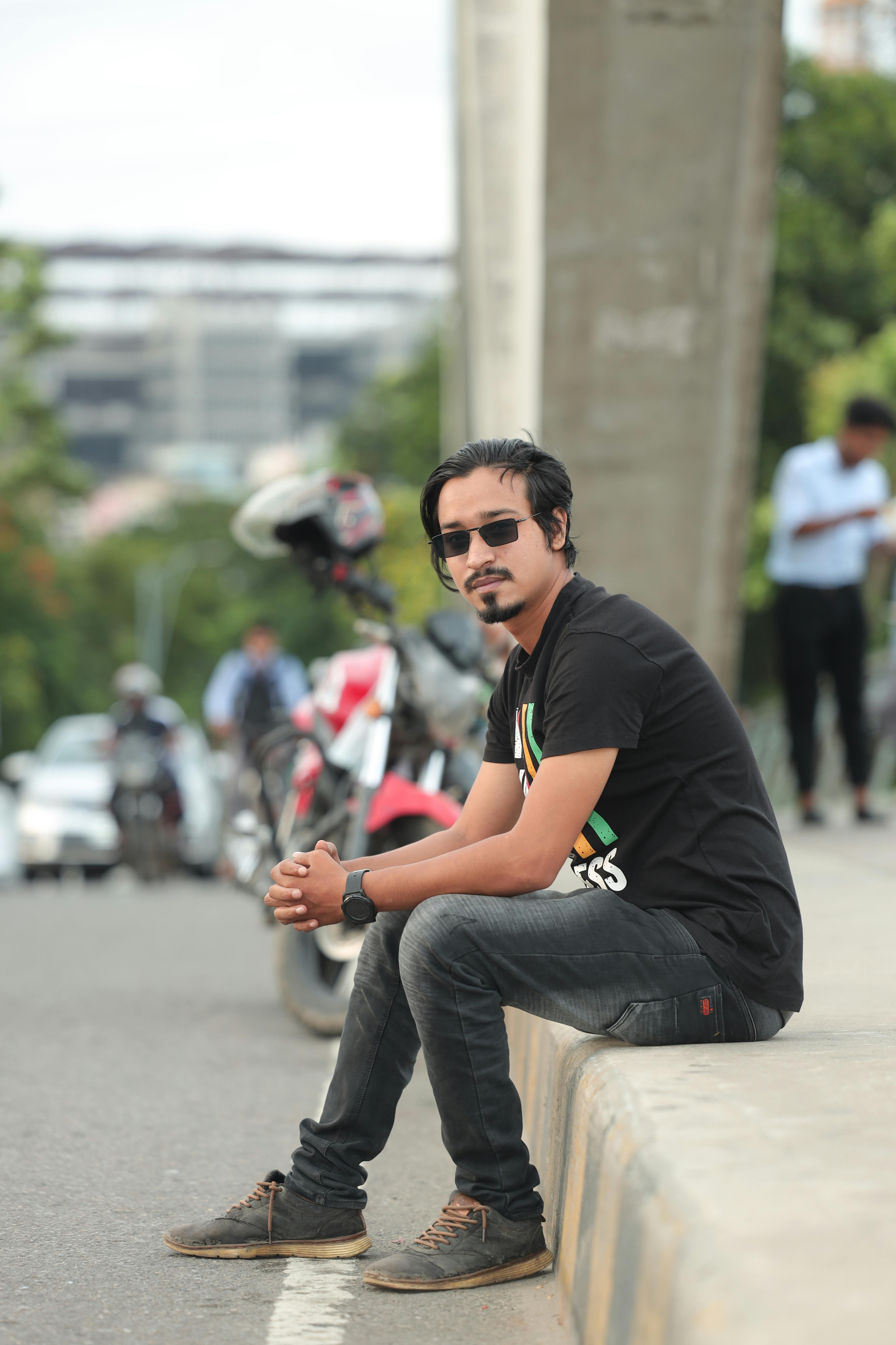 Casual portrait of a man sitting on a Dhaka sidewalk wearing sunglasses and a black shirt.