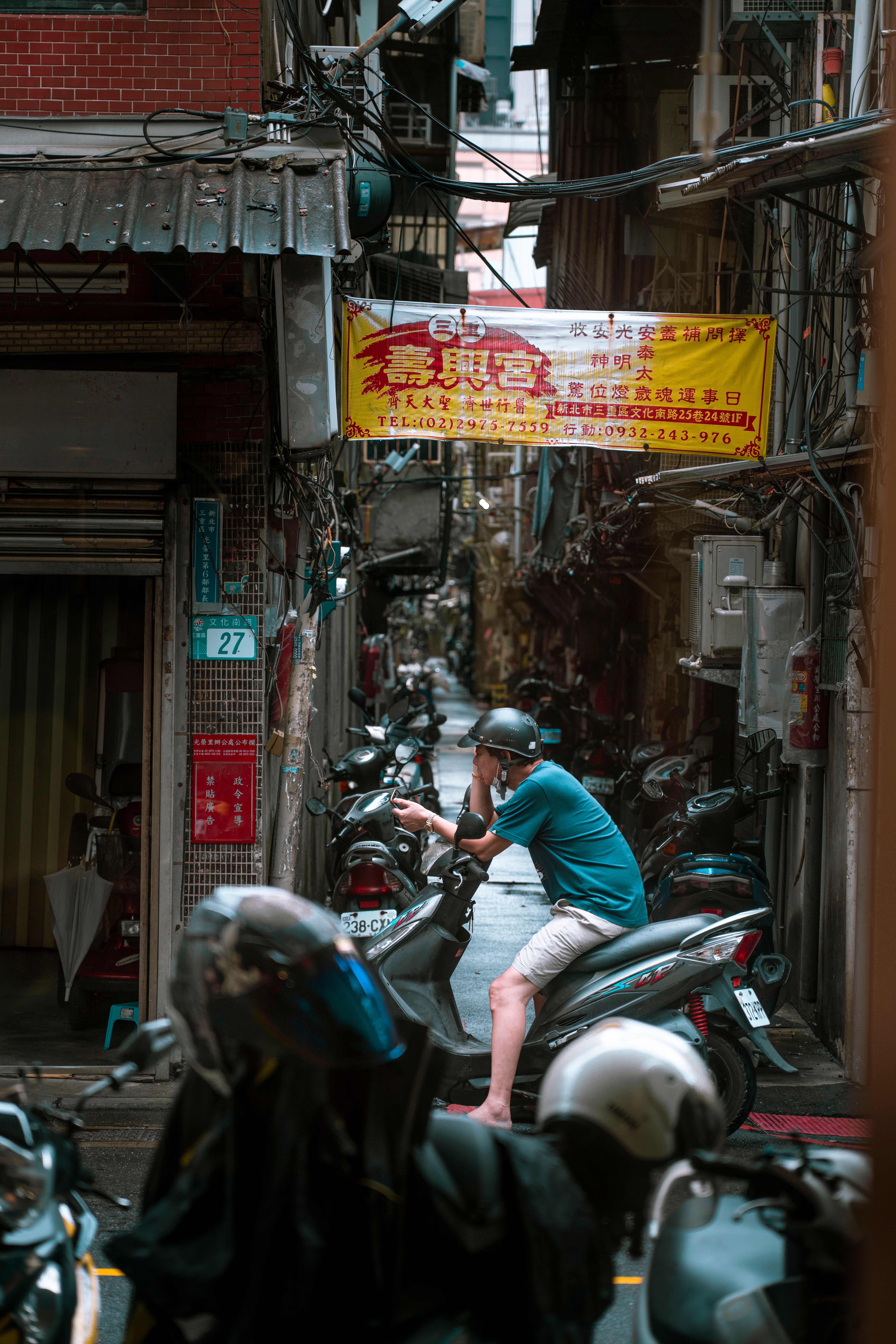 A man on a scooter in a narrow urban alley filled with parked motorbikes.