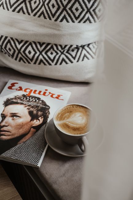 A cappuccino with latte art next to a stylish magazine and cushion.